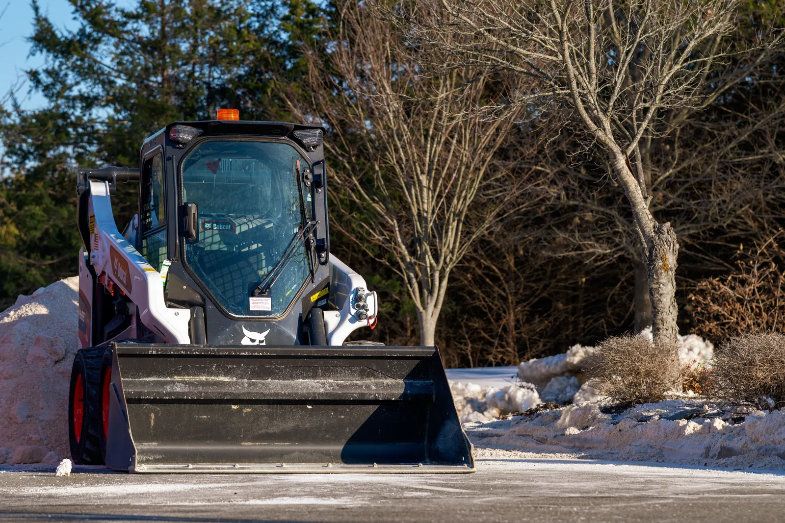 Skid-steer loader fitted with a snow attachment clearing a paved commercial lot on a clear winter morning